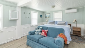 A bedroom with a bed, a blue sofa, a wooden nightstand, and a window with blinds. - Clamdigger PETS  Ocean Block. Screened Porch (Tybee Island)