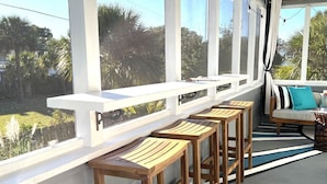 A sunlit outdoor seating area with wooden bar stools and a cushioned bench. - Clamdigger PETS  Ocean Block. Screened Porch (Tybee Island)