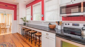 A kitchen with white cabinets, a black countertop, and a red accent wall. There are three bar stools, a wooden bowl, and a kettle on the stove. - Clamdigger PETS  Ocean Block. Screened Porch (Tybee Island)