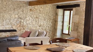 A rustic room with a stone wall, a wooden dining table with woven placemats, a leather sofa, and a window with a view of greenery.
