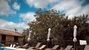 Une piscine entourée d’arbres et d’un bâtiment dont le toit est recouvert de tuiles, avec des chaises longues et des parasols à proximité.