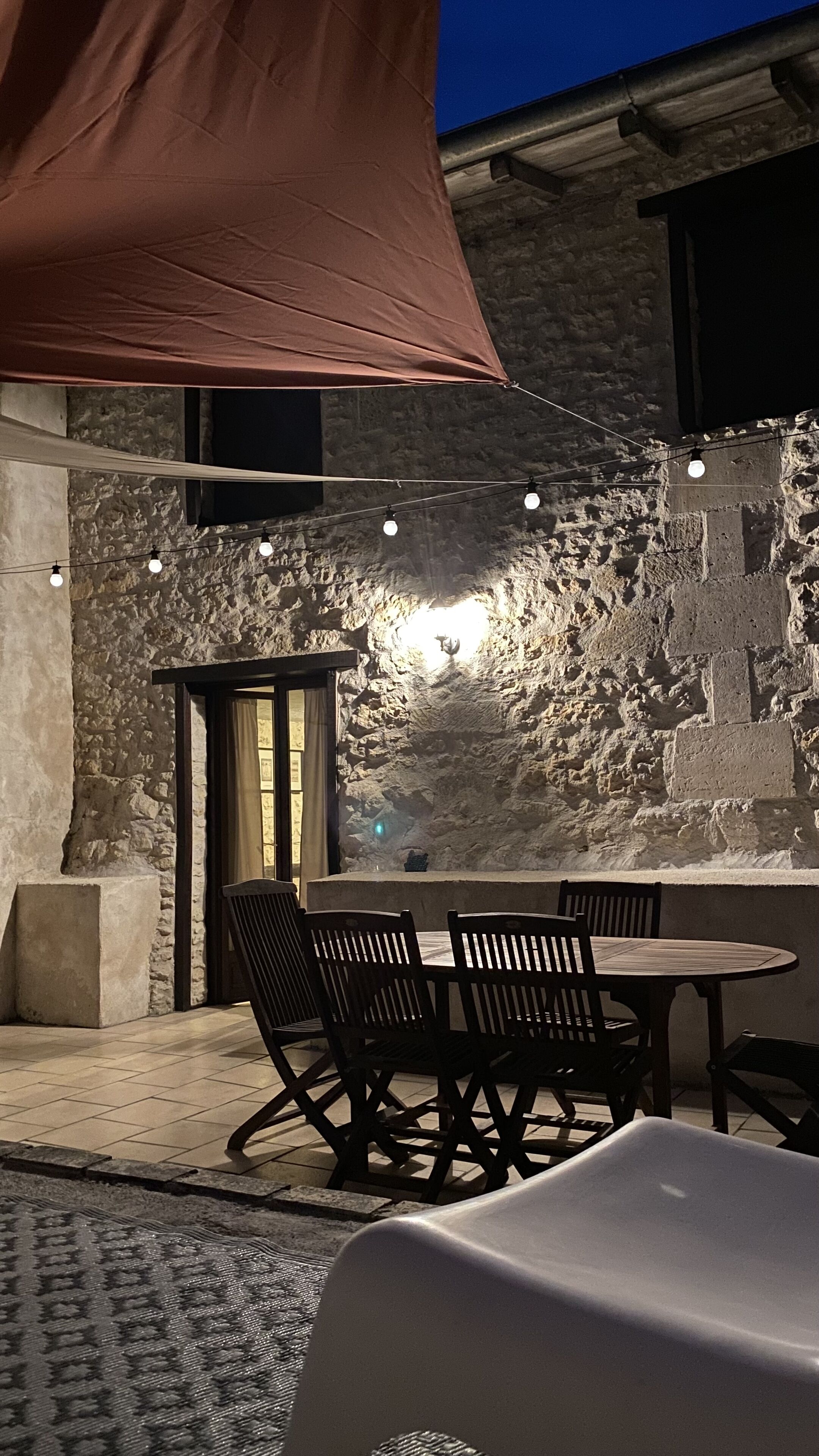 An outdoor dining area with stone walls, a round table, and chairs. There is a canopy overhead and string lights.