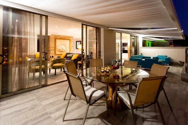A modern dining area with a glass table and chairs, a stone accent wall, and a cozy seating area with sofas and cushions. - Penthouse with great terrace and sea views (Mogán)