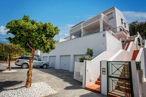 Un bâtiment moderne de couleur blanche, avec une voiture garée devant, un arbre et une entrée fermée par un portail.