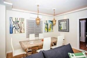 A dining area with a wooden table, white chairs, and two hanging chandeliers. There are abstract paintings on the walls.