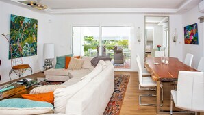 Un salon spacieux avec un canapé blanc, une table à manger en bois et un grand miroir. Un balcon, sur lequel on aperçoit une chaise et une table, est accessible par des portes vitrées.