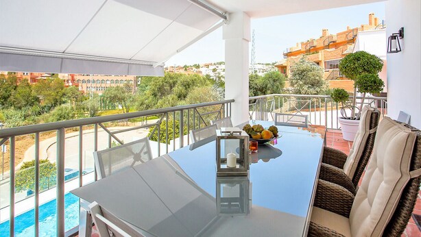 Un balcon avec une table en verre, des chaises en osier, un pot de fleurs et une vue sur le paysage urbain.