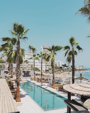 Ein Poolbereich mit Palmen, Liegestühlen und Blick auf Strand und Meer.