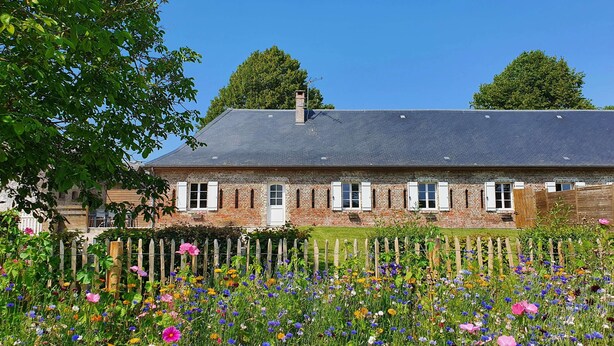A brick house with a sloped roof and white shutters, surrounded by a garden with various flowers.