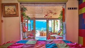 A bedroom with a colorful quilt, a painting on the wall, and a view of the outdoors.