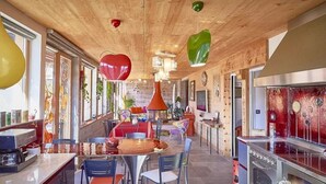 A restaurant with a wooden ceiling, a central dining area with round tables and chairs, and a kitchen area with stainless steel appliances.
