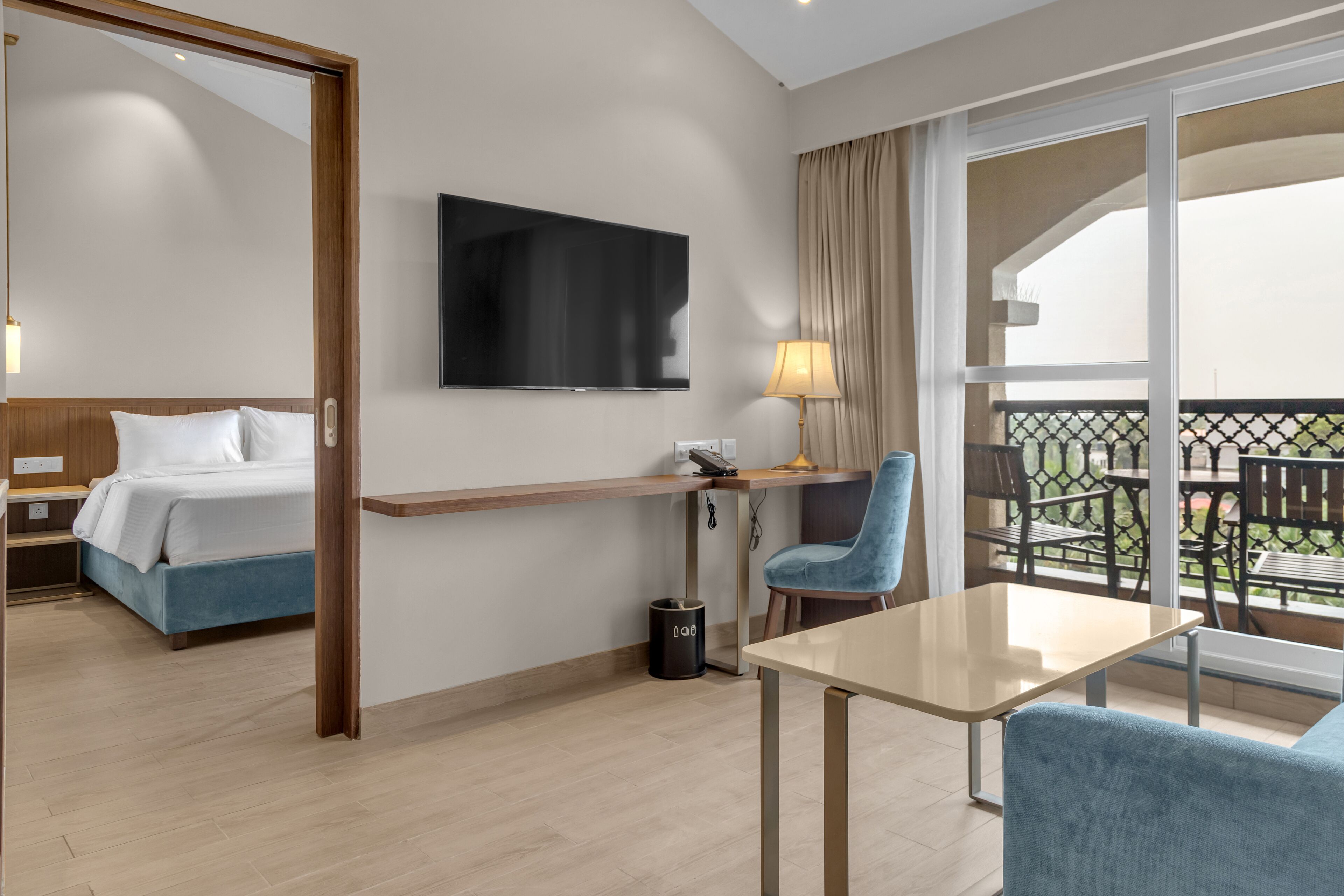 A hotel room with a bed, a desk, a chair, a TV, and a balcony with outdoor furniture.