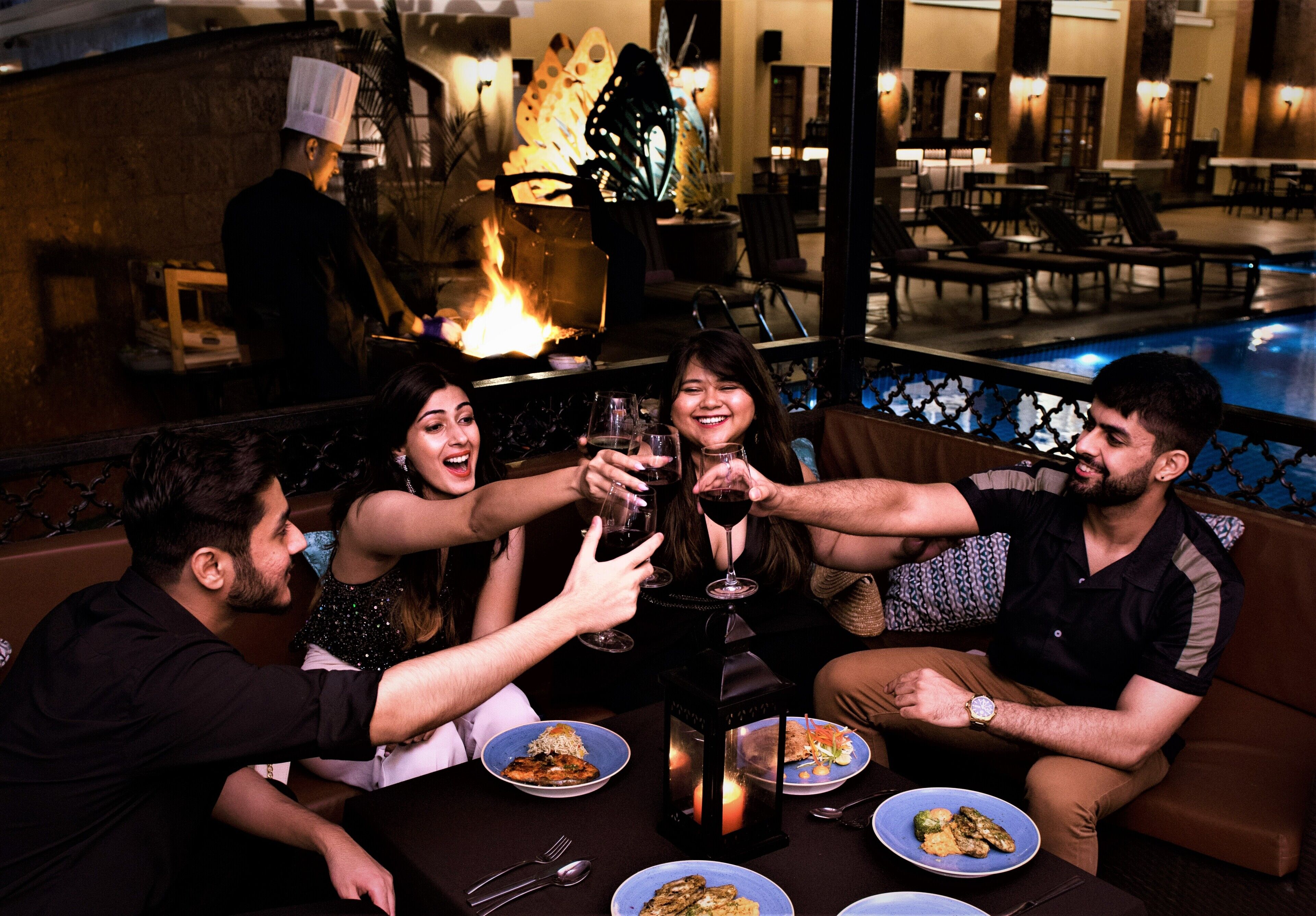 Un groupe de personnes trinquant autour d’une table où sont disposés des plats de nourriture, une lanterne allumée et une piscine en arrière-plan.