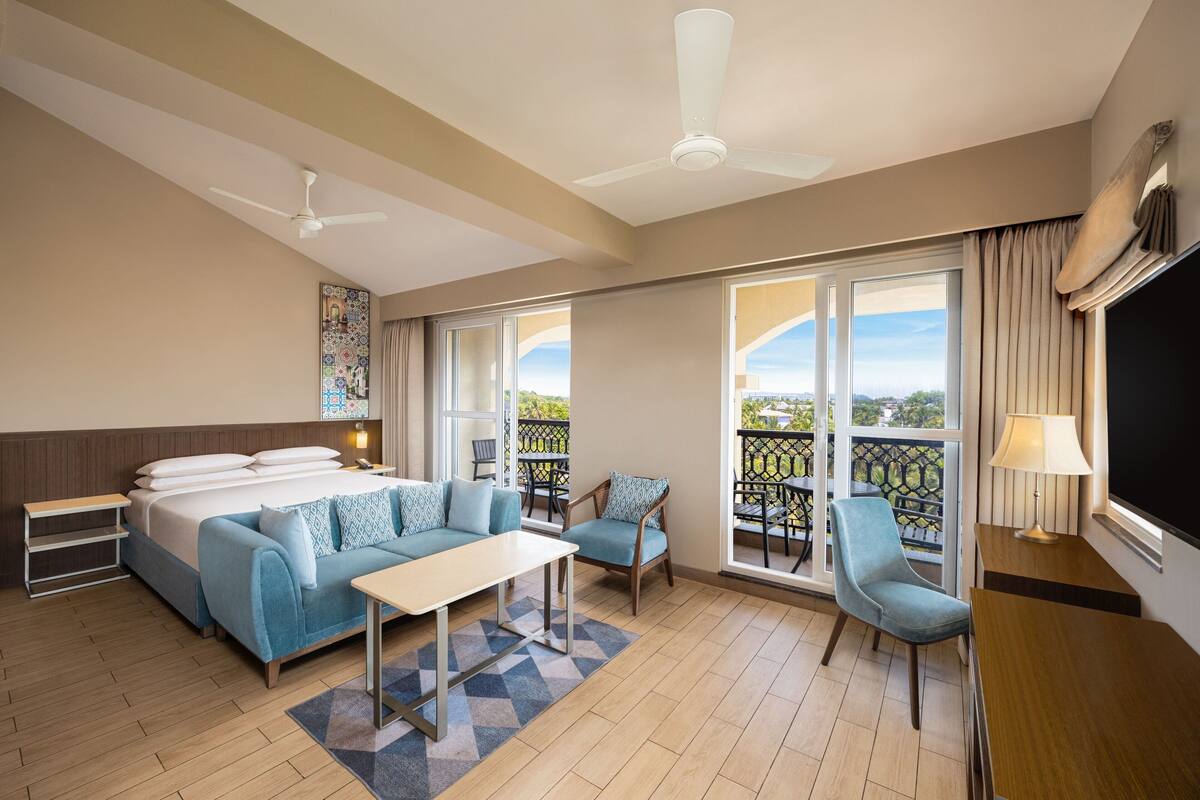 Habitación de hotel con cama, sofá, mesa, sillas, balcón con vistas y ventilador de techo.