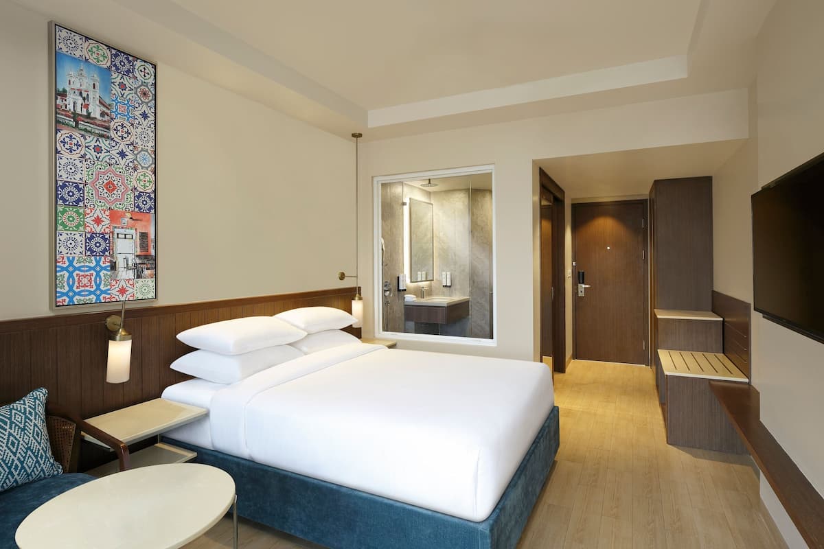Habitación de hotel con una cama grande, una mesita de noche, una silla, un televisor y un baño visible a través de una puerta abierta.