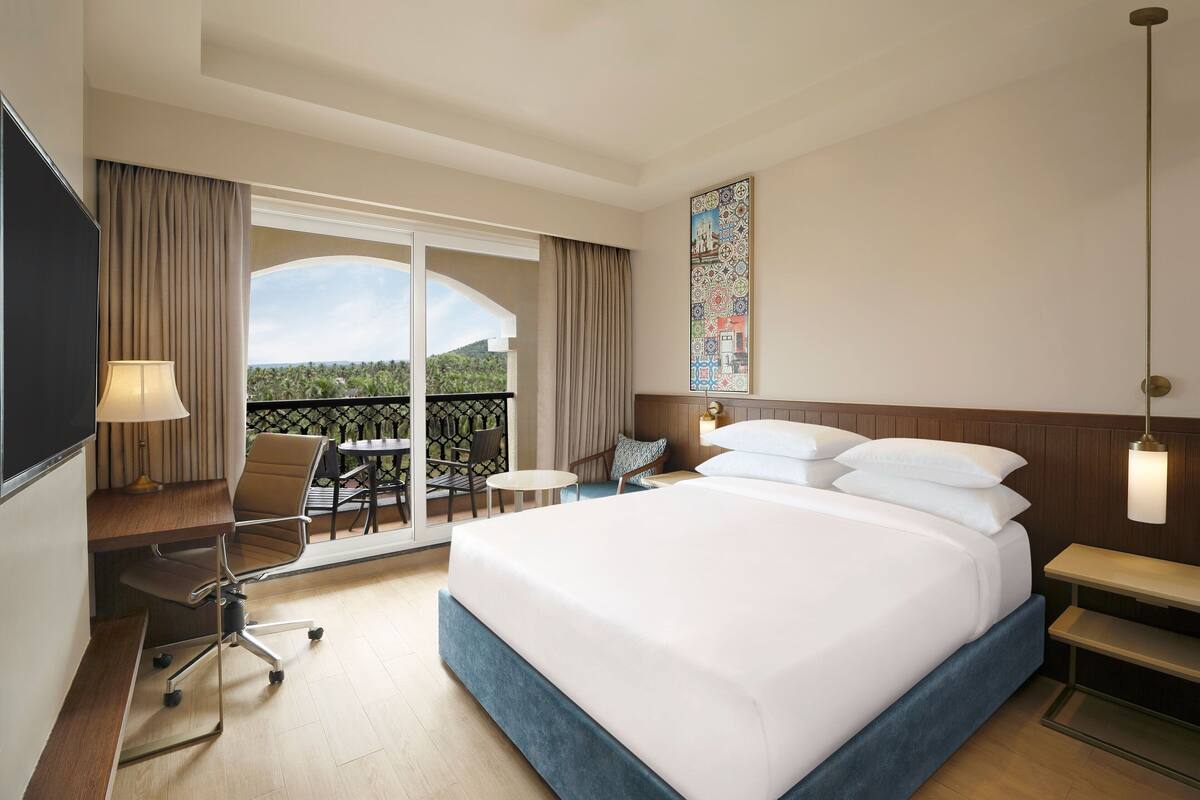 Habitación de hotel con una cama grande, un escritorio con silla, una mesita y un balcón con vistas al paisaje.
