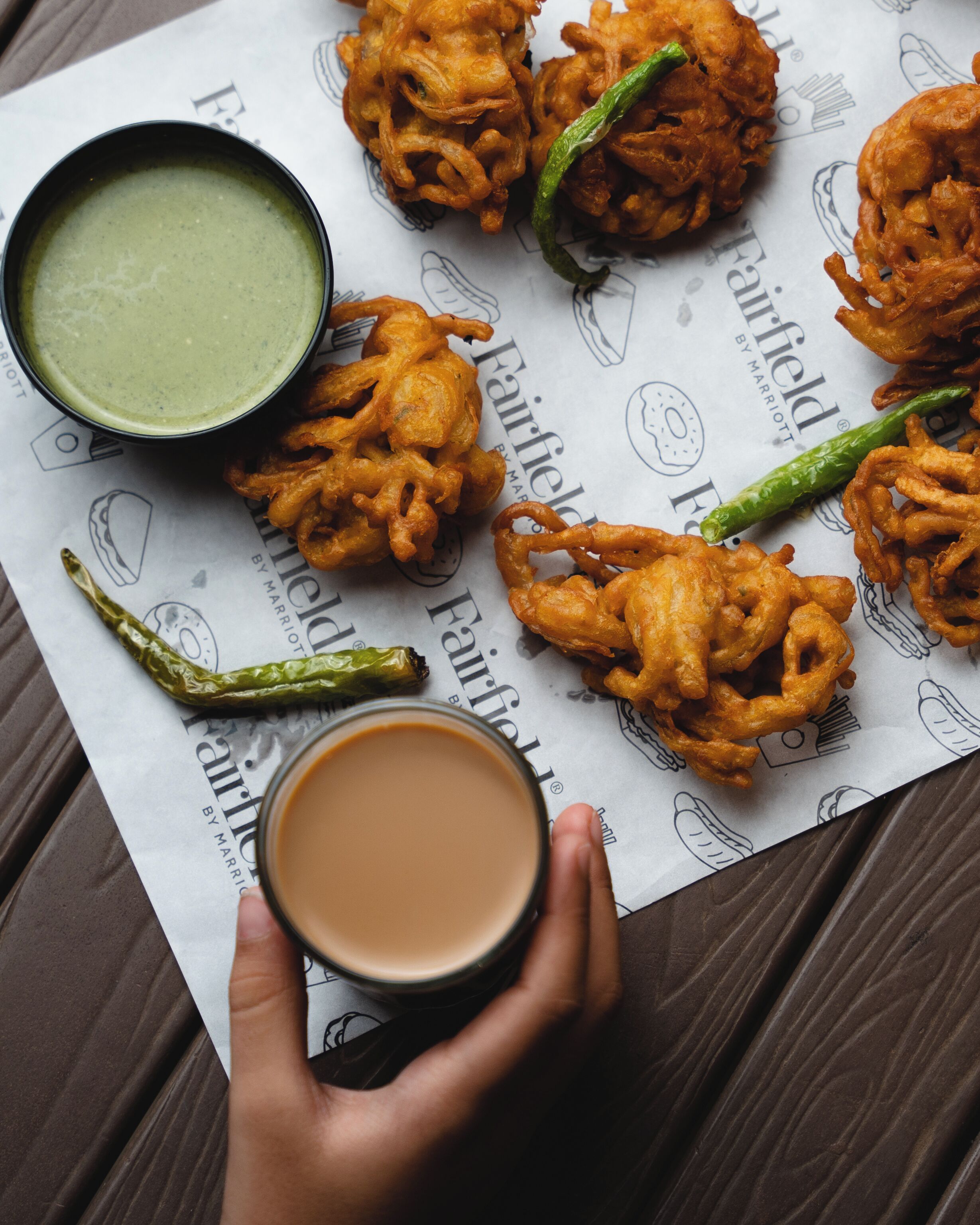 A tray of fried snacks, a bowl of green sauce, a green chili, and a cup of tea on a table with a patterned paper.