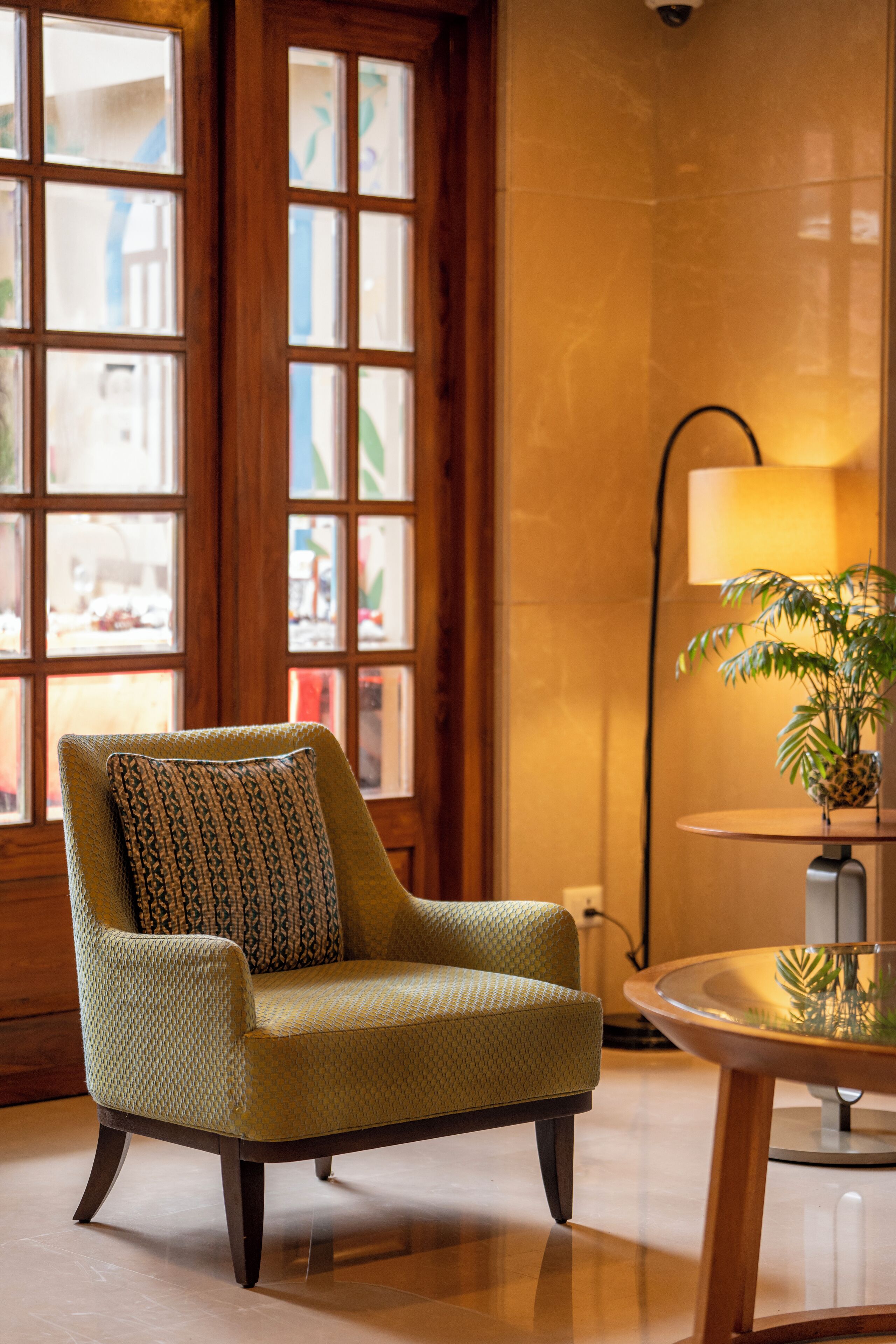 A room with a green armchair, a round table with a lamp, and a window with a view of buildings.