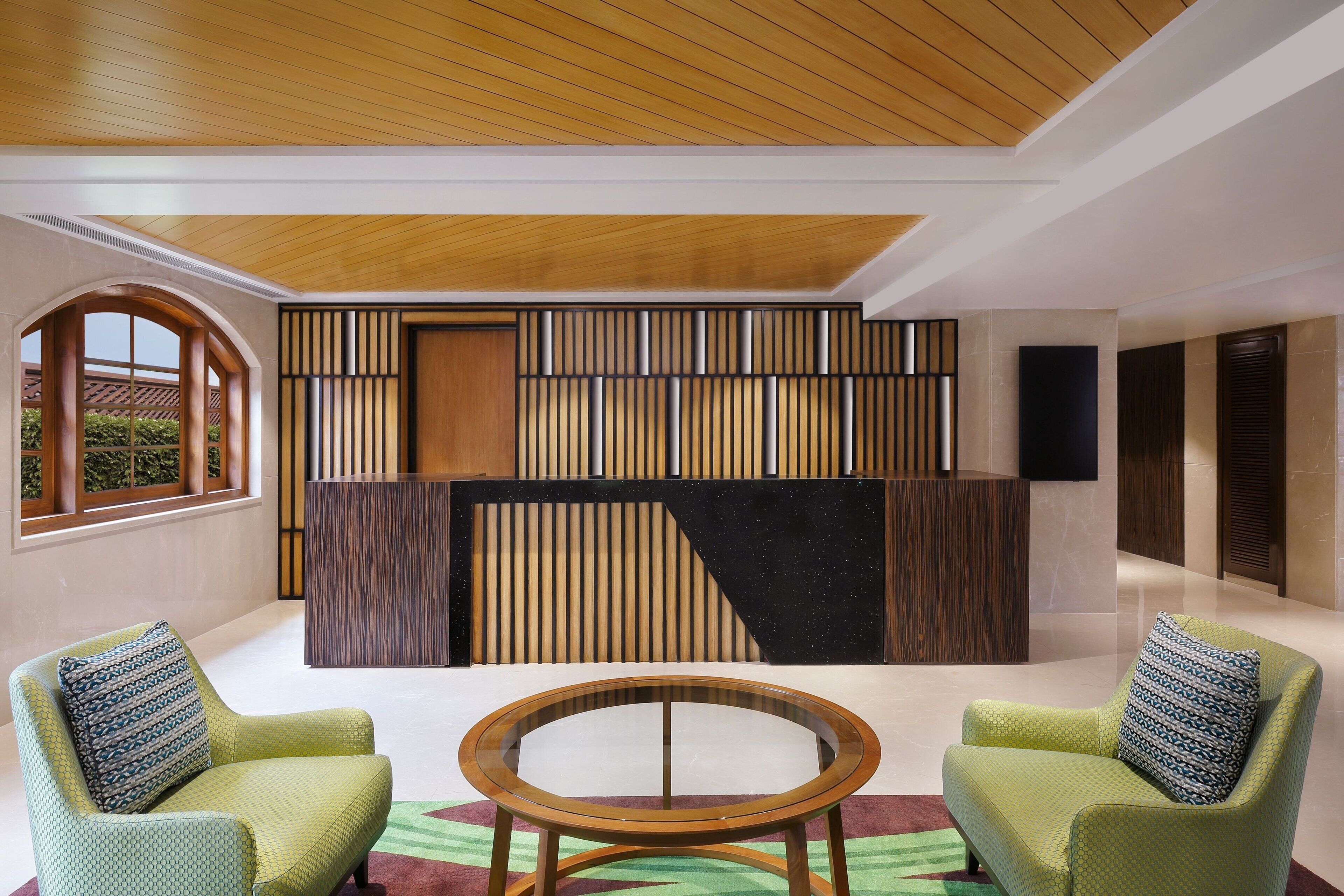 A modern lobby with a wooden ceiling, a reception desk, and two green armchairs with patterned cushions.