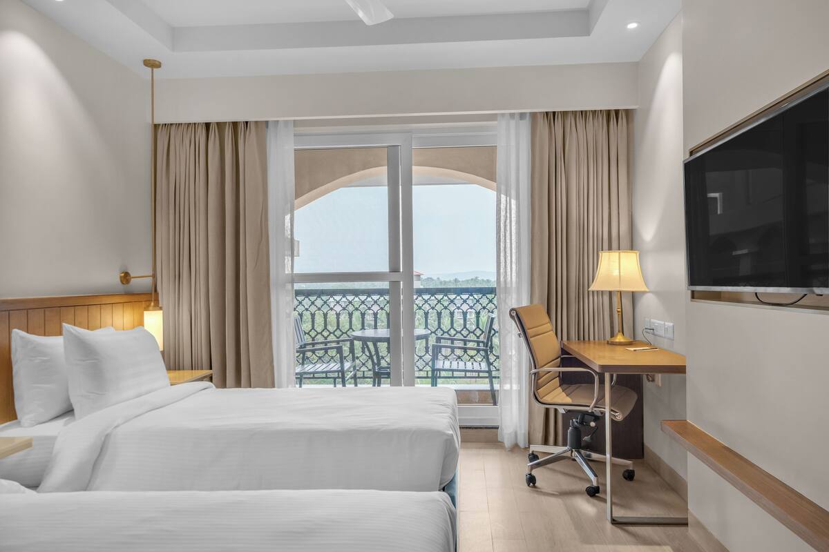 Habitación de hotel con cama, escritorio con silla, televisión y balcón con vistas.