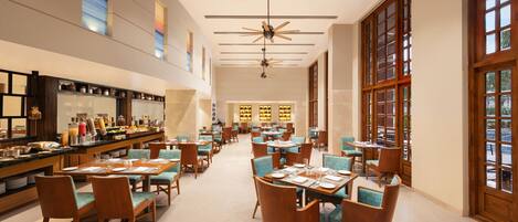 Un vaste espace de restauration dans l’hôtel, avec de nombreuses tables et chaises, de grandes fenêtres et des ventilateurs de plafond.