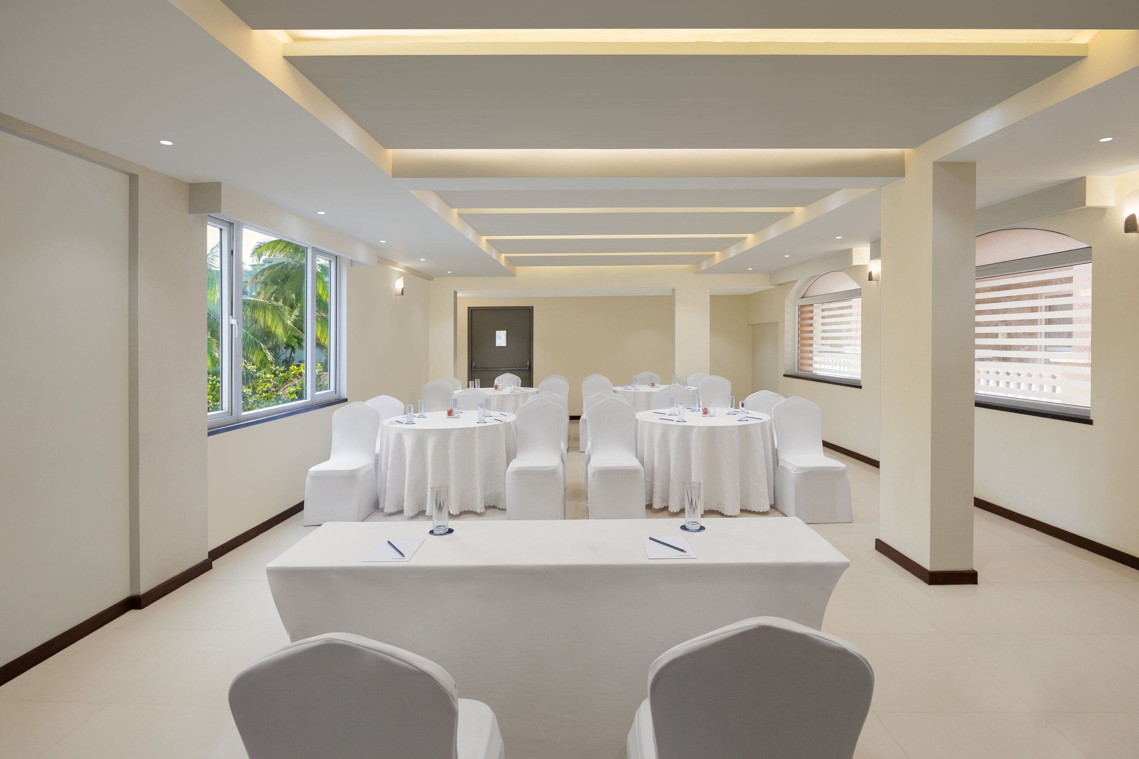 Un salón de conferencias con mesas y sillas blancas, ventanas amplias y un diseño de techo moderno.
