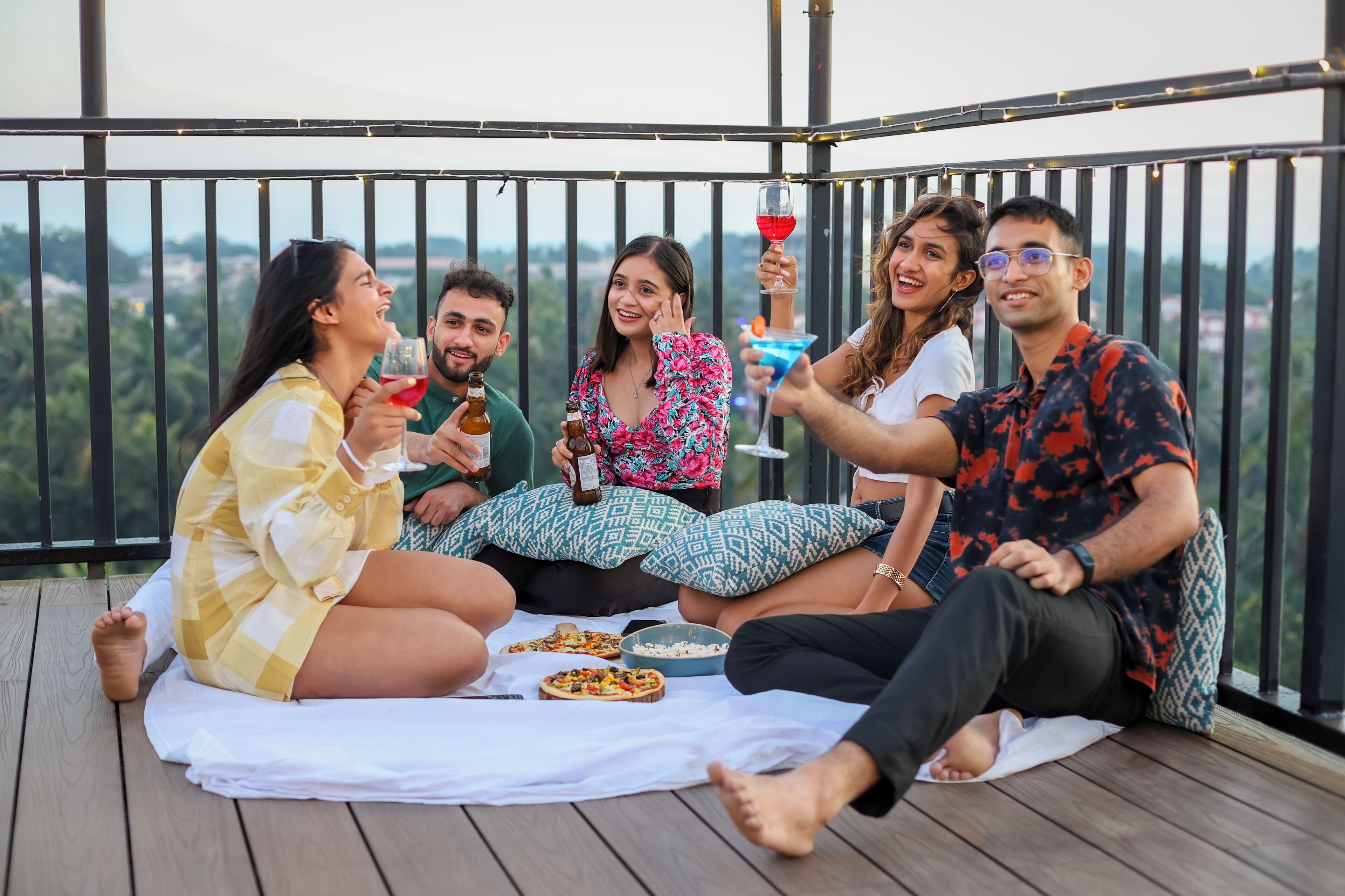 A group of people sitting on a wooden deck with a white tablecloth, enjoying drinks and pizza.