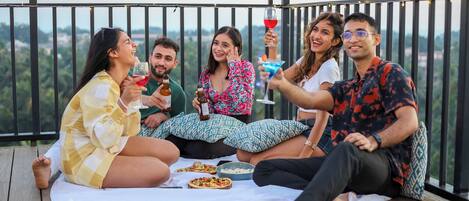 Un grupo de personas sentadas en una terraza de madera con mantel blanco, disfrutando de bebidas y pizza.