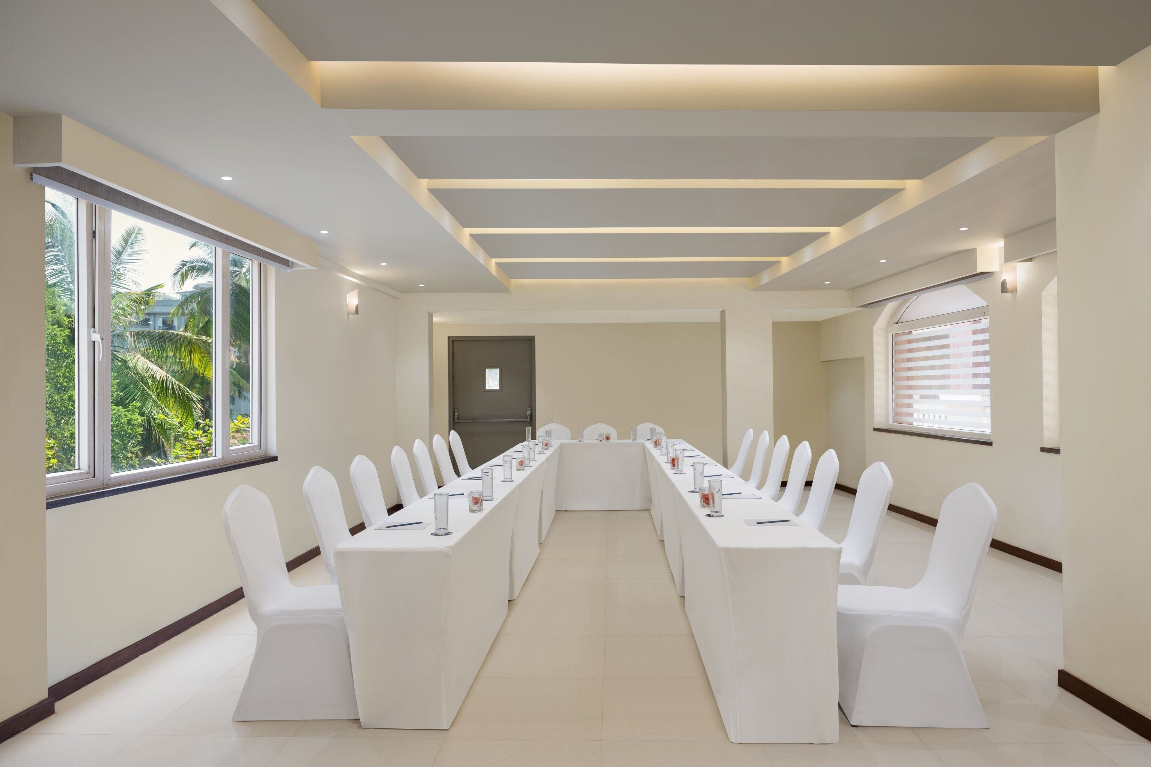 A conference room with a long table, white chairs, and large windows with a view of greenery.