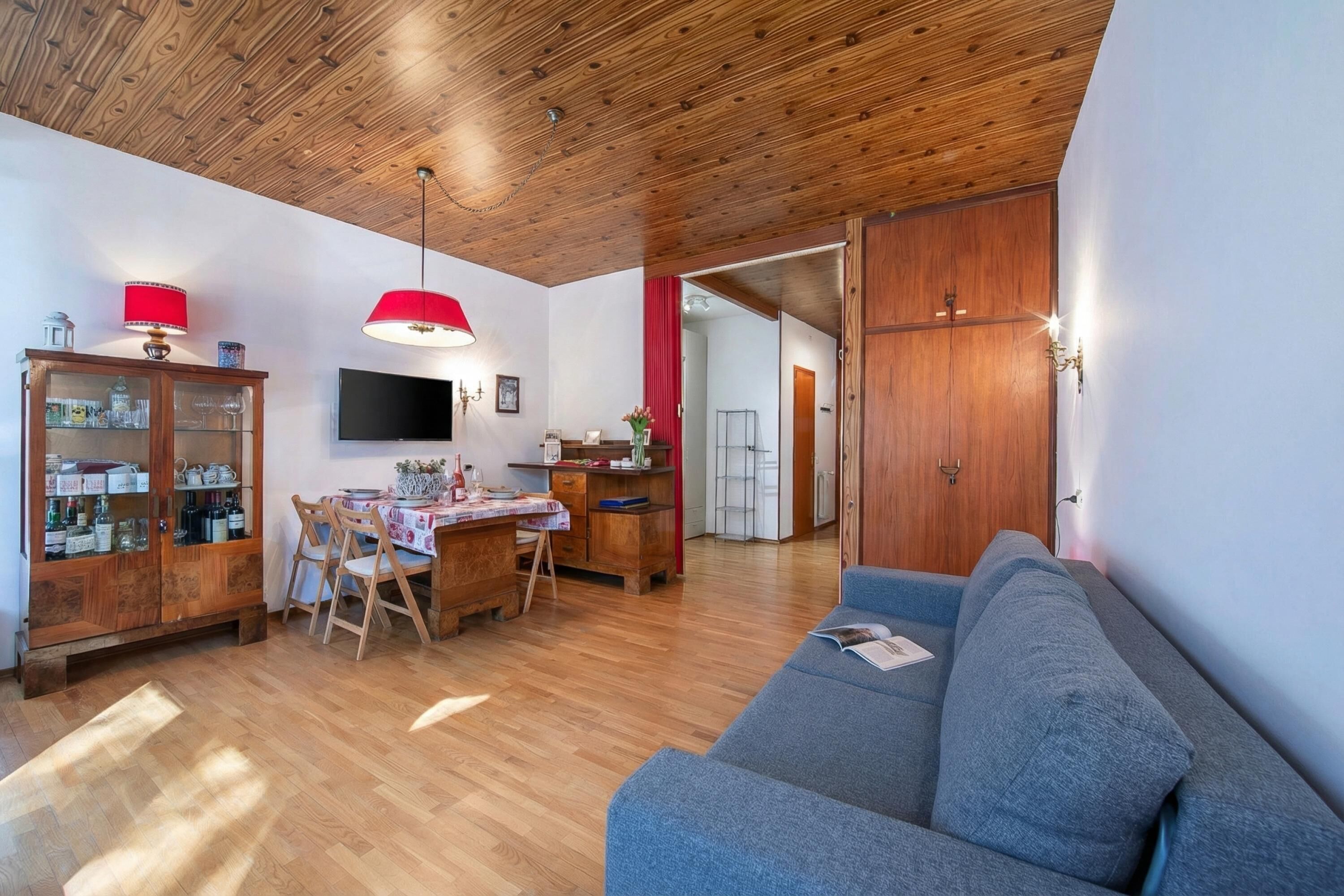 Ein Wohnzimmer mit Holzboden, einem blauen Sofa, einem Esstisch mit Stühlen, einem Fernseher und einer Vitrine mit Glastüren.