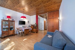 Een woonkamer met een houten plafond, een blauwe bank, een eettafel met stoelen, een televisie en een kast met glazen deuren.