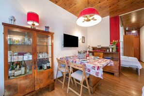 Een eetgedeelte met een houten tafel voor vier personen, een kast met glazen deuren, een aan de muur bevestigde televisie en een bed met een rood gordijn.