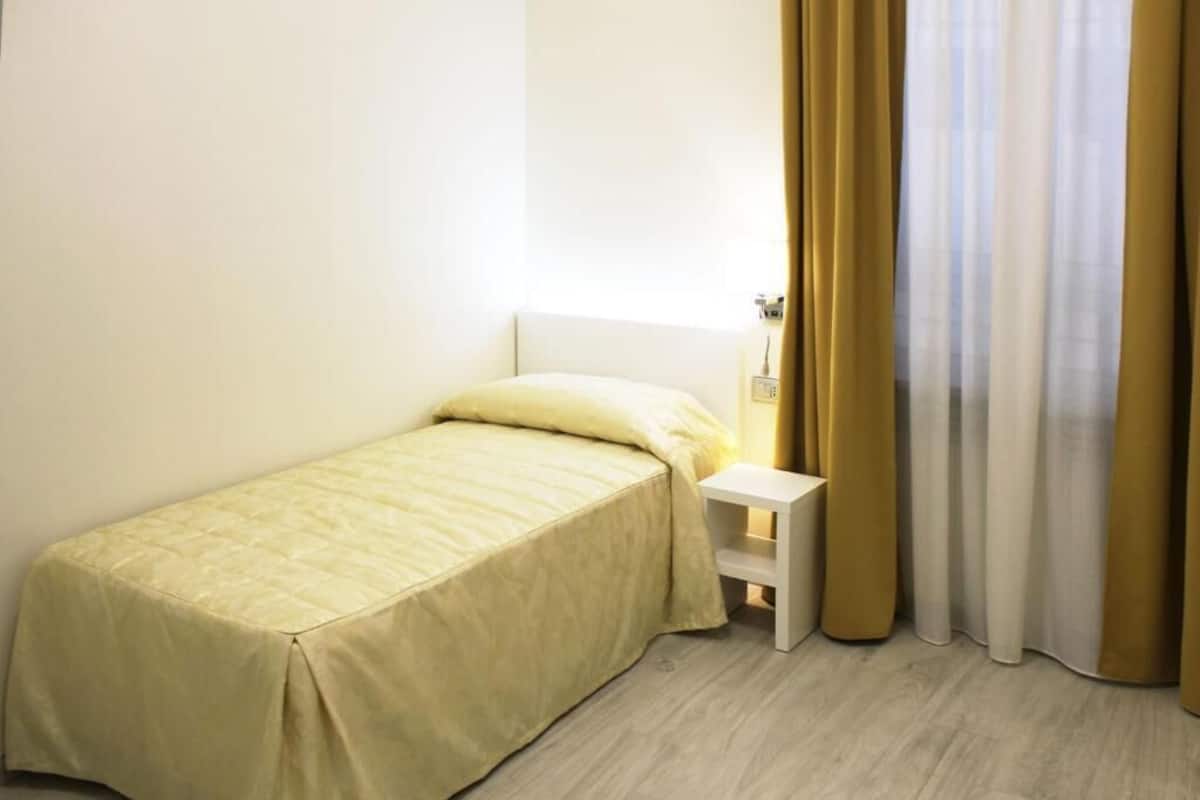 Habitación pequeña con cama individual, cabecero blanco, mesita de noche y ventana con cortinas color beige.