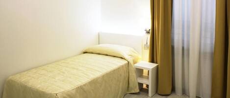 Habitación pequeña con cama individual, cabecero blanco, mesita de noche y ventana con cortinas color beige.