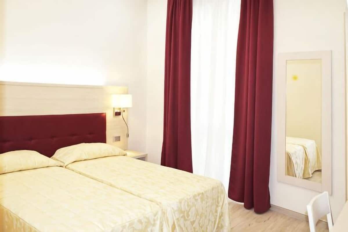 Habitación de hotel con cama, mesita de noche, espejo y cortinas rojas.