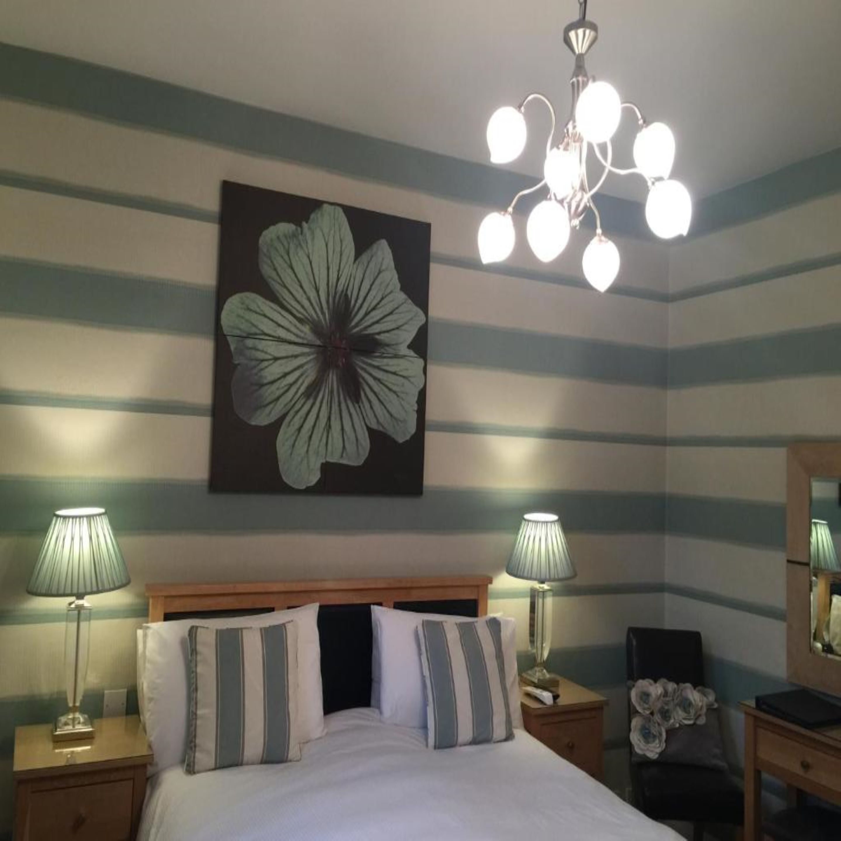 A hotel room with a bed, two bedside tables, a nightstand, a chair, a mirror, and a chandelier.