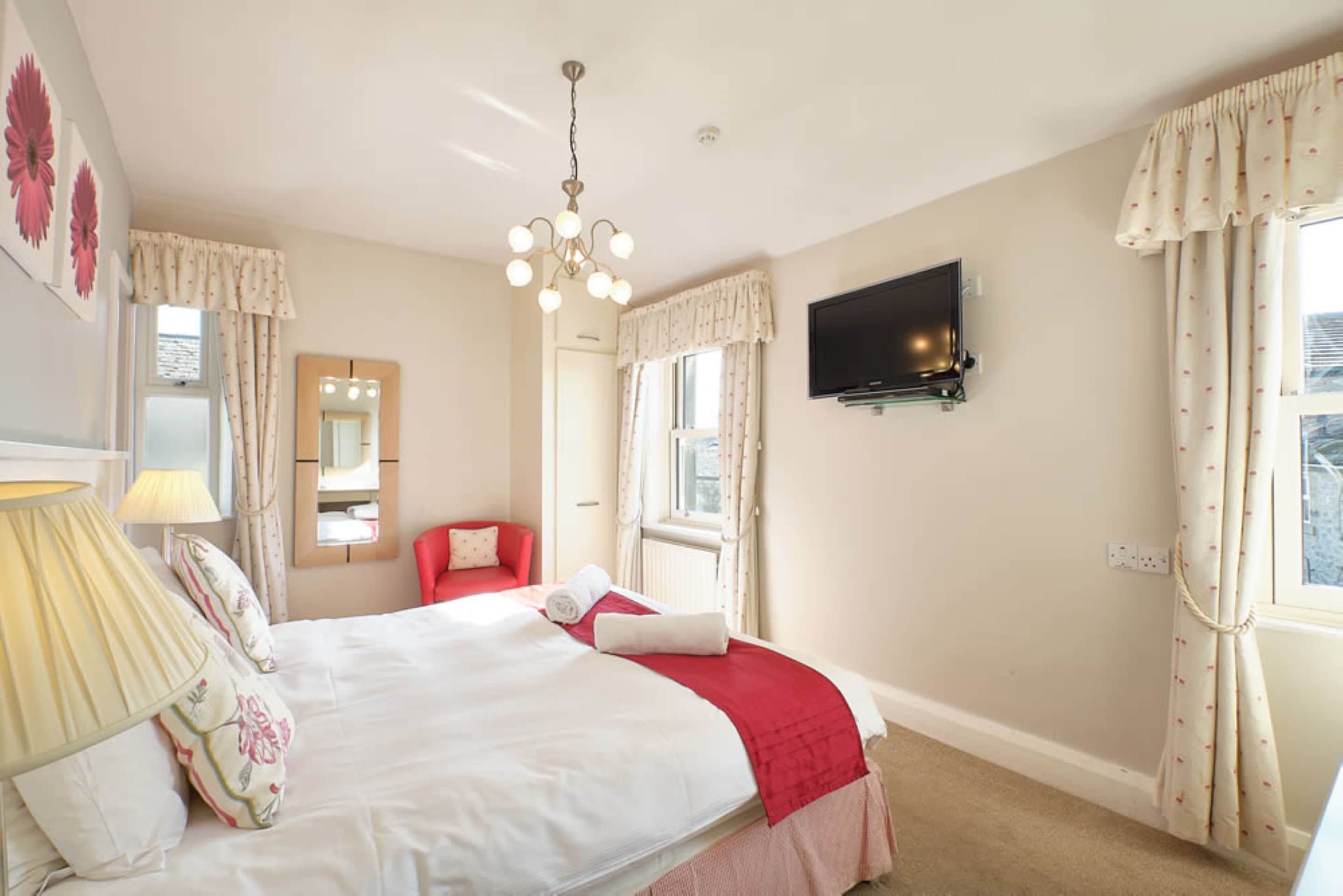 A hotel room with a bed, a TV mounted on the wall, a window with curtains, a chair, and a lamp.