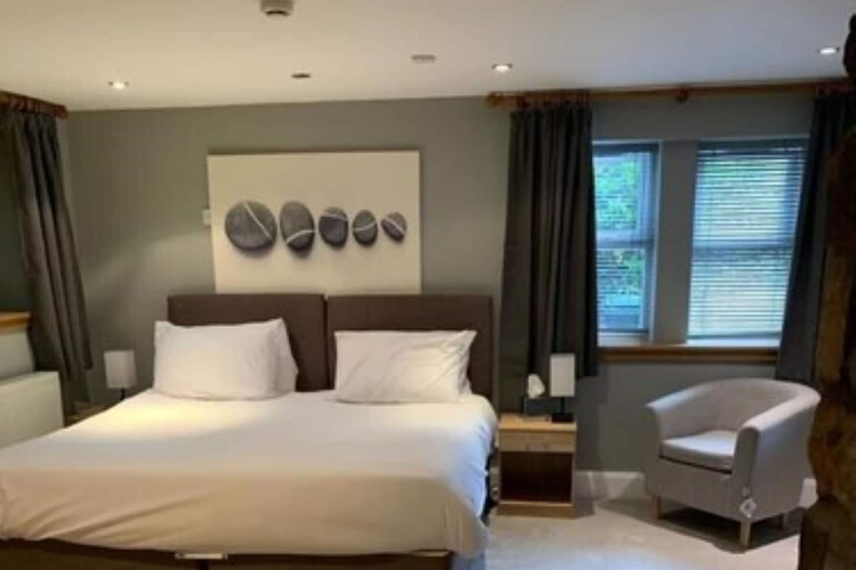 A bedroom with a large bed, a nightstand, a chair, and a stone wall.