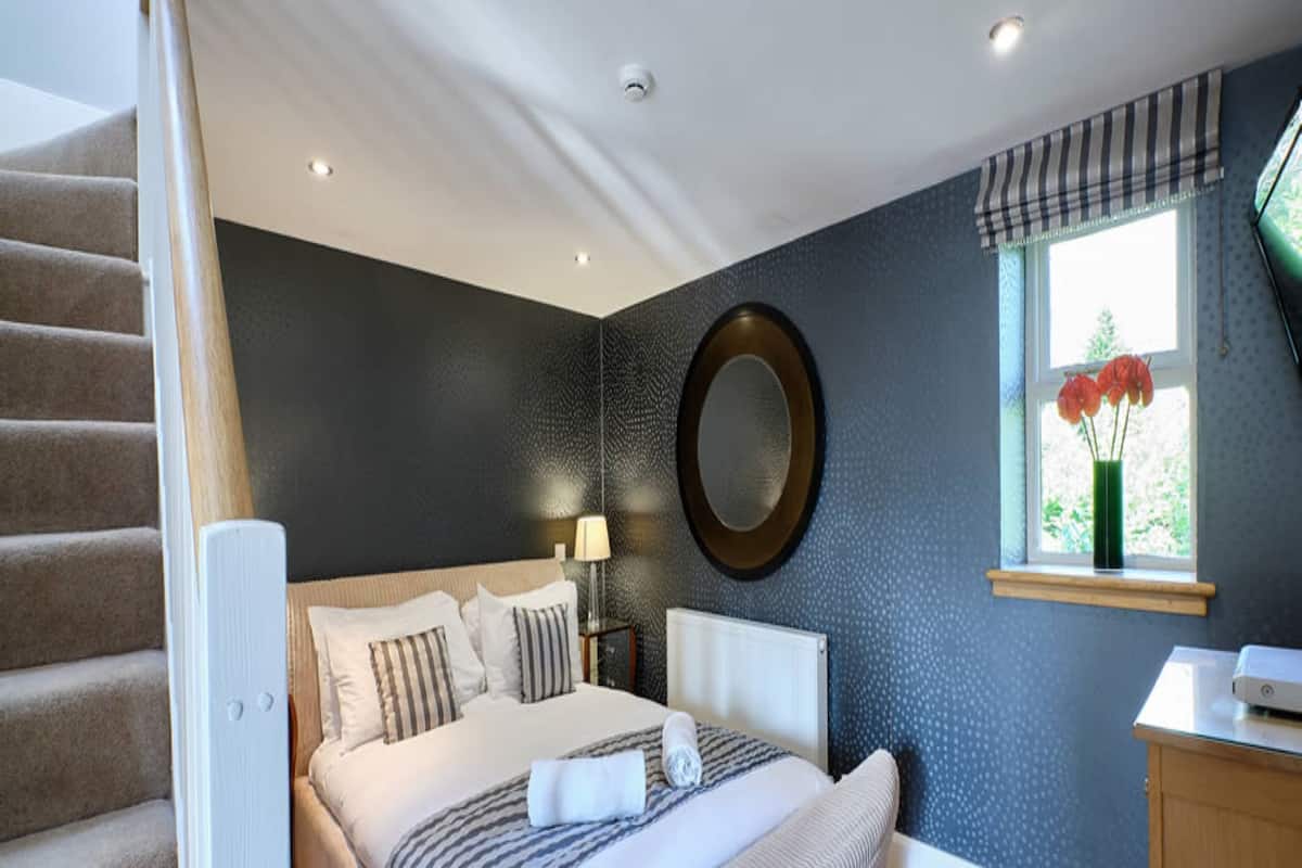A bedroom with a bed, a window with flowers, a lamp, and a round mirror on the wall.