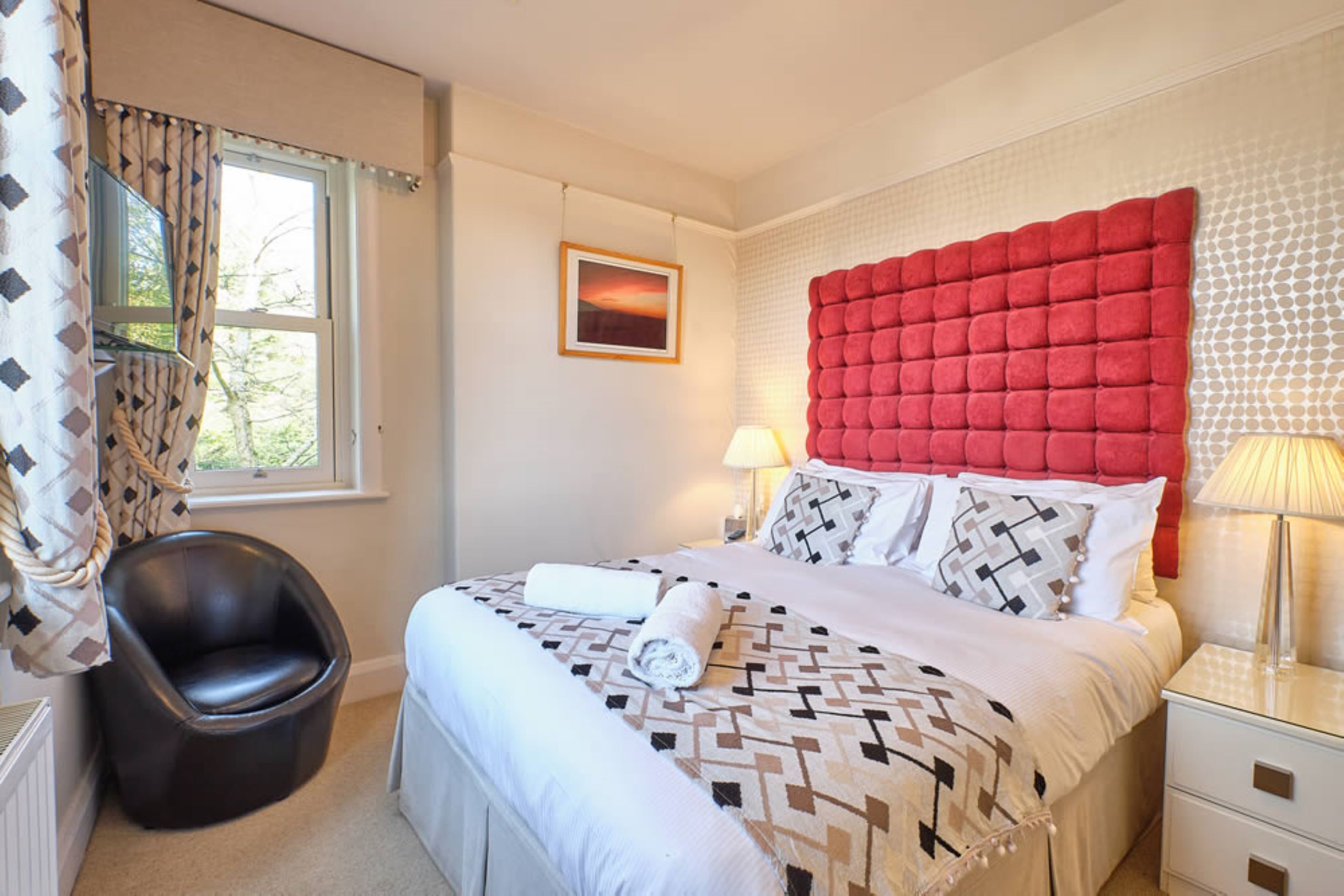 Une chambre d’hôtel avec un grand lit, une tête de lit, une table de chevet avec une lampe, une chaise et une fenêtre avec des rideaux.