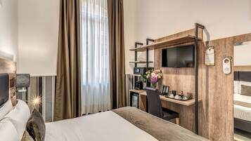 A hotel room with a bed, a desk with a computer, a TV, and a small table with a flower vase.