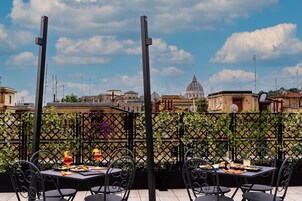 Zona pranzo all'aperto con tavoli e sedie, una recinzione decorativa e vista su un edificio storico sullo sfondo.