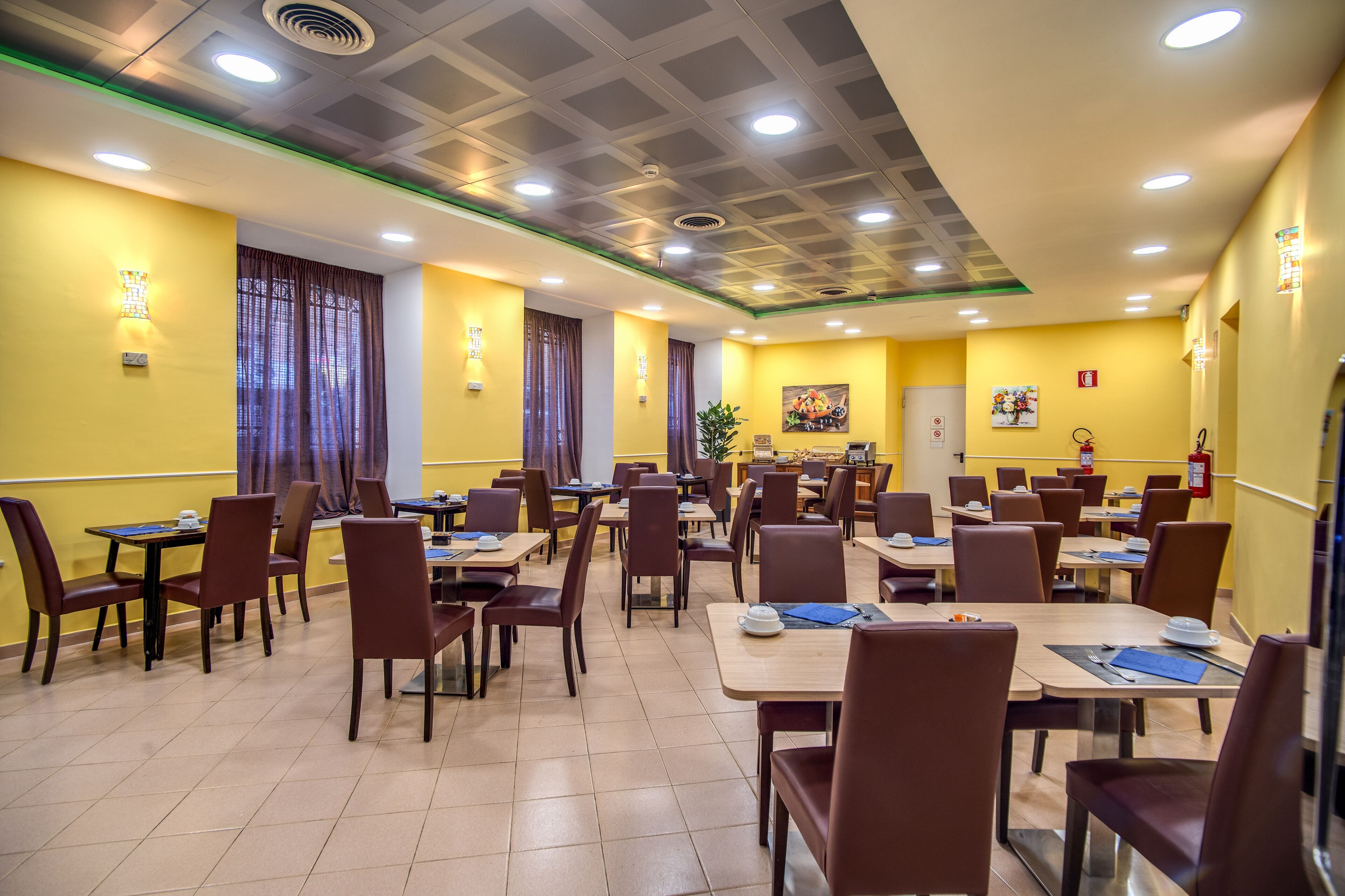 A dining area with tables and chairs, yellow walls, and ceiling lights.