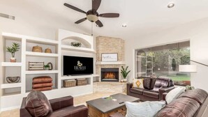 A modern living room with a fireplace, leather sofas, a flat-screen TV, and a ceiling fan. - Dreaming Tree Retreat | Pool, Spa & Fire Pit (Phoenix)