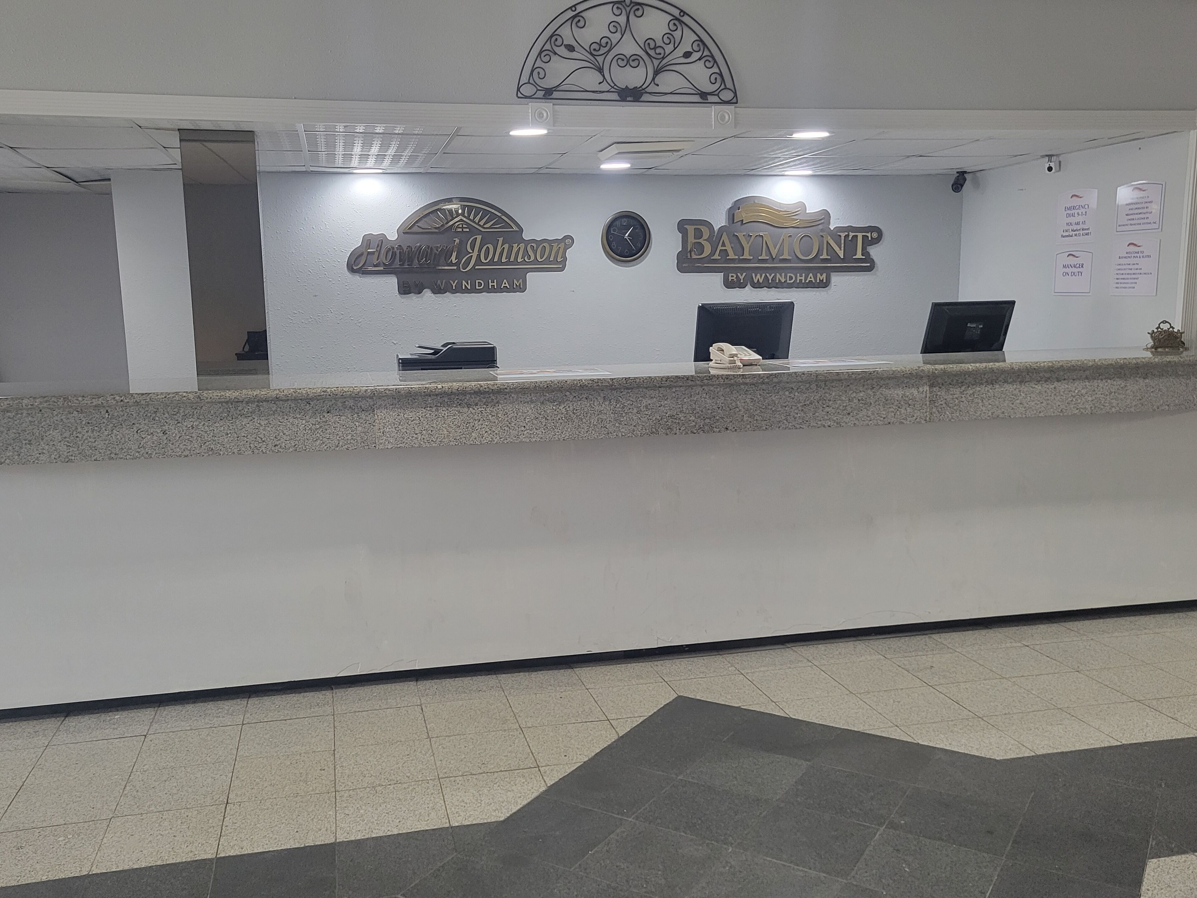 Un comptoir d’accueil avec une horloge, un ordinateur et un téléphone. On peut voir des panneaux indiquant « Howard Johnson » et « Baymont » sur le mur.