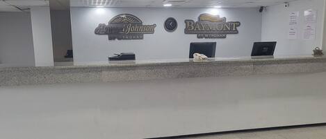 Un comptoir d’accueil avec une horloge, un ordinateur et un téléphone. On peut voir des panneaux indiquant « Howard Johnson » et « Baymont » sur le mur.