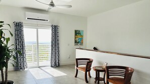 Quarto com ventoinha no teto, ar condicionado, duas cadeiras de madeira, uma mesinha, um vaso de planta, janela com cortinas estampadas e um quadro na parede.