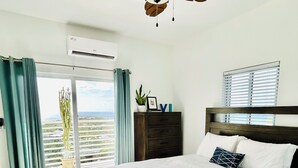 Quarto com cama grande, ventilador de teto, cômoda e vista pela janela.