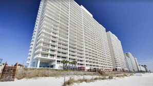 Ein Hochhaus-Hotel mit mehreren Balkonen, nahe einem Strand mit sandigem Vordergrund und blauem, wolkenlosem Himmel.