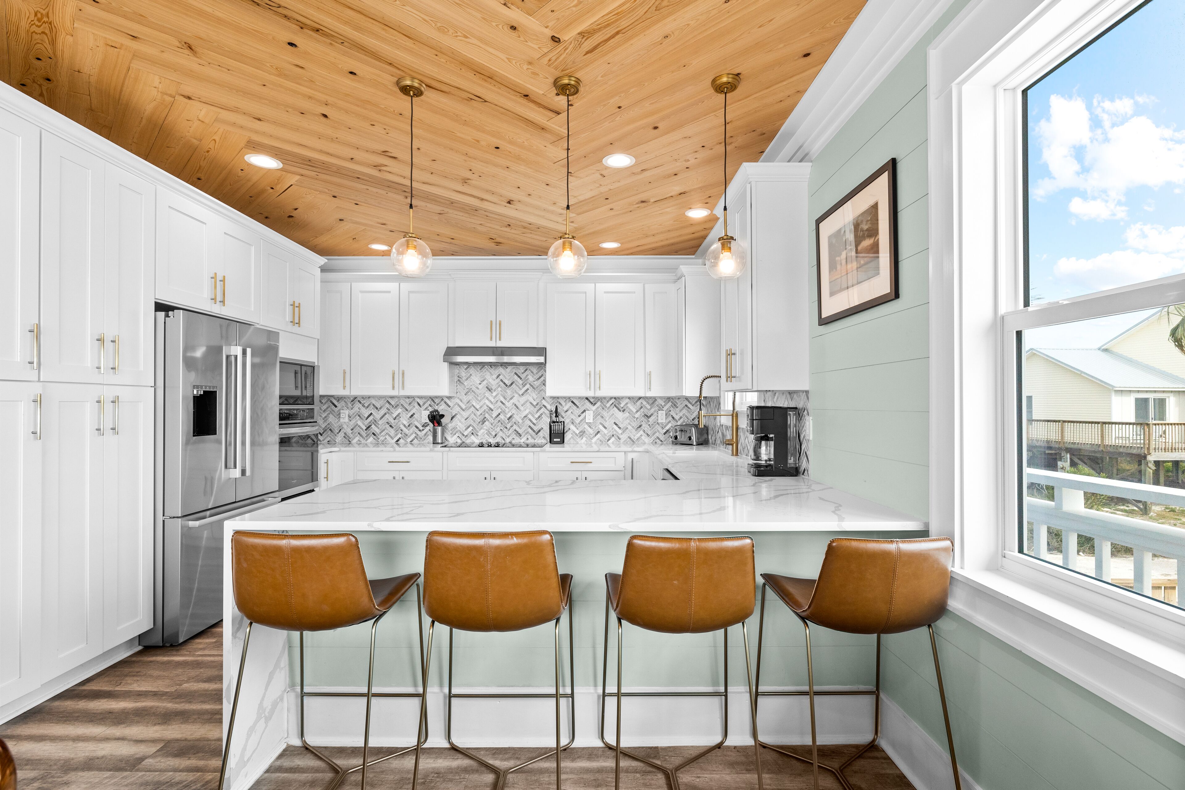 A modern kitchen with white cabinets, a large island with bar stools, a stainless steel refrigerator, and a wooden ceiling.